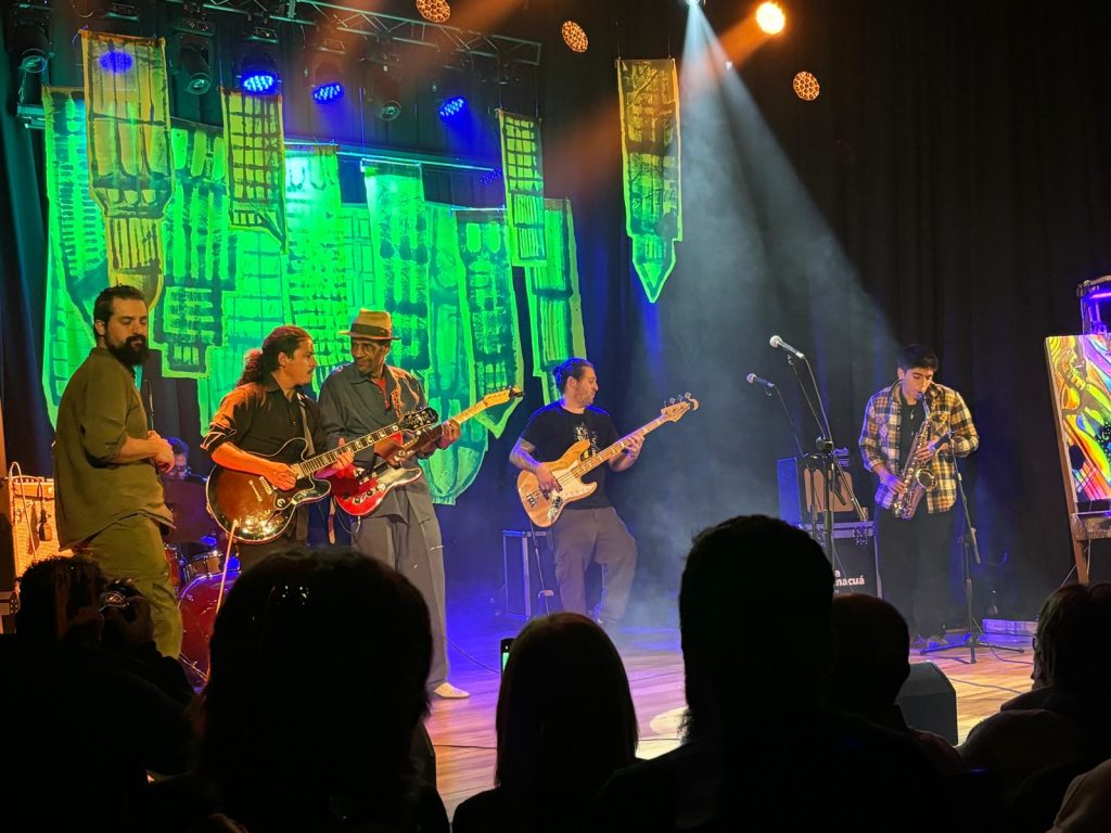 El pasado martes 23/07 en Sala Camacuá se produjo el octavo concierto organizado por el Club del Blues en una interesante iniciativa que consiste en traer a consagrados y veteranos artistas del blues norteamericano para tocar con una banda soporte local. En la previa se presentaron en dúo Clara Lassus y Junior Flores, y luego La Máquina a Vapor como teloneros, haciendo clásicos con sus dinámicos y acostumbrados cambios de instrumentistas y artistas sobre el escenario. Nunca decepcionan. El escenario lució renovado telón y la iluminación estuvo a cargo de Guillermo Ifrán. Con entradas agotadas en una sala colmada de público multigeneracional (hasta familiar) y también inclusivo, se apersonó Michael Dotson presentado como "el blues de Chicago que ya no se toca en Chicago", soportado por Adrián Flores en batería, Hernán Poloni en guitarra y Junior Flores en bajo. Ante una audiencia rendida a sus pies, tocó y cantó durante más de una hora, generando momentos de tensa intimidad y también picos de euforia. No soy cronista musical por lo tanto no voy a tener el atrevimiento de comentar en ese aspecto, pero el show claramente fue muy satisfactorio para el público que festejaba cada comentario (volaban las traducciones simultáneas susurradas), al mismo tiempo que a un costado del escenario un colectivo de artistas plásticos iba generando una obra pictórica en tiempo real, con el buen tino de incorporar pintura fluorescente, adecuadamente iluminada. Finalizó con bises sumando a los artistas locales, a toda fiesta. Un gran concierto en suma, evento multisensorial, en una sala cómoda, accesible, con baños renovados y expendio de bebidas que se pueden llevar a la sala para acompañar debidamente el espectáculo con la promesa de luego retornar los vasos y colaborar con la limpieza. Evento que ya no es el secreto mejor guardado de la escena musical montevideana, como evidenciaba la larga cola al ingreso. Sigan al Club del Blues en sus redes para enterarse del próximo show, que será en setiembre y habrán anticipadas bonificadas. El pasado martes 23/07 en Sala Camacuá se produjo el octavo concierto organizado por el Club del Blues en una interesante iniciativa que consiste en traer a consagrados y veteranos artistas del blues norteamericano para tocar con una banda soporte local.En la previa se presentaron en dúo Clara Lassus y Junior Flores, y luego La Máquina a Vapor como teloneros, haciendo clásicos con sus dinámicos y acostumbrados cambios de instrumentistas y artistas sobre el escenario. Nunca decepcionan.
El escenario lució renovado telón y la iluminación estuvo a cargo de Guillermo Ifrán.
Con entradas agotadas en una sala colmada de público multigeneracional (hasta familiar) y también inclusivo, se apersonó Michael Dotson presentado como "el blues de Chicago que ya no se toca en Chicago", soportado por Adrián Flores en batería, Hernán Poloni en guitarra y Junior Flores en bajo.
Ante una audiencia rendida a sus pies, tocó y cantó durante más de una hora, generando momentos de tensa intimidad y también picos de euforia.
No soy cronista musical por lo tanto no voy a tener el atrevimiento de comentar en ese aspecto, pero el show claramente fue muy satisfactorio para el público que festejaba cada comentario (volaban las traducciones simultáneas susurradas), al mismo tiempo que a un costado del escenario un colectivo de artistas plásticos iba generando una obra pictórica en tiempo real, con el buen tino de incorporar pintura fluorescente, adecuadamente iluminada.
Finalizó con bises sumando a los artistas locales, a toda fiesta.
Un gran concierto en suma, evento multisensorial, en una sala cómoda, accesible, con baños renovados y expendio de bebidas que se pueden llevar a la sala para acompañar debidamente el espectáculo con la promesa de luego retornar los vasos y colaborar con la limpieza.
Evento que ya no es el secreto mejor guardado de la escena musical montevideana, como evidenciaba la larga cola al ingreso.
Sigan al Club del Blues en sus redes para enterarse del próximo show, que será en setiembre y habrán anticipadas bonificadas.